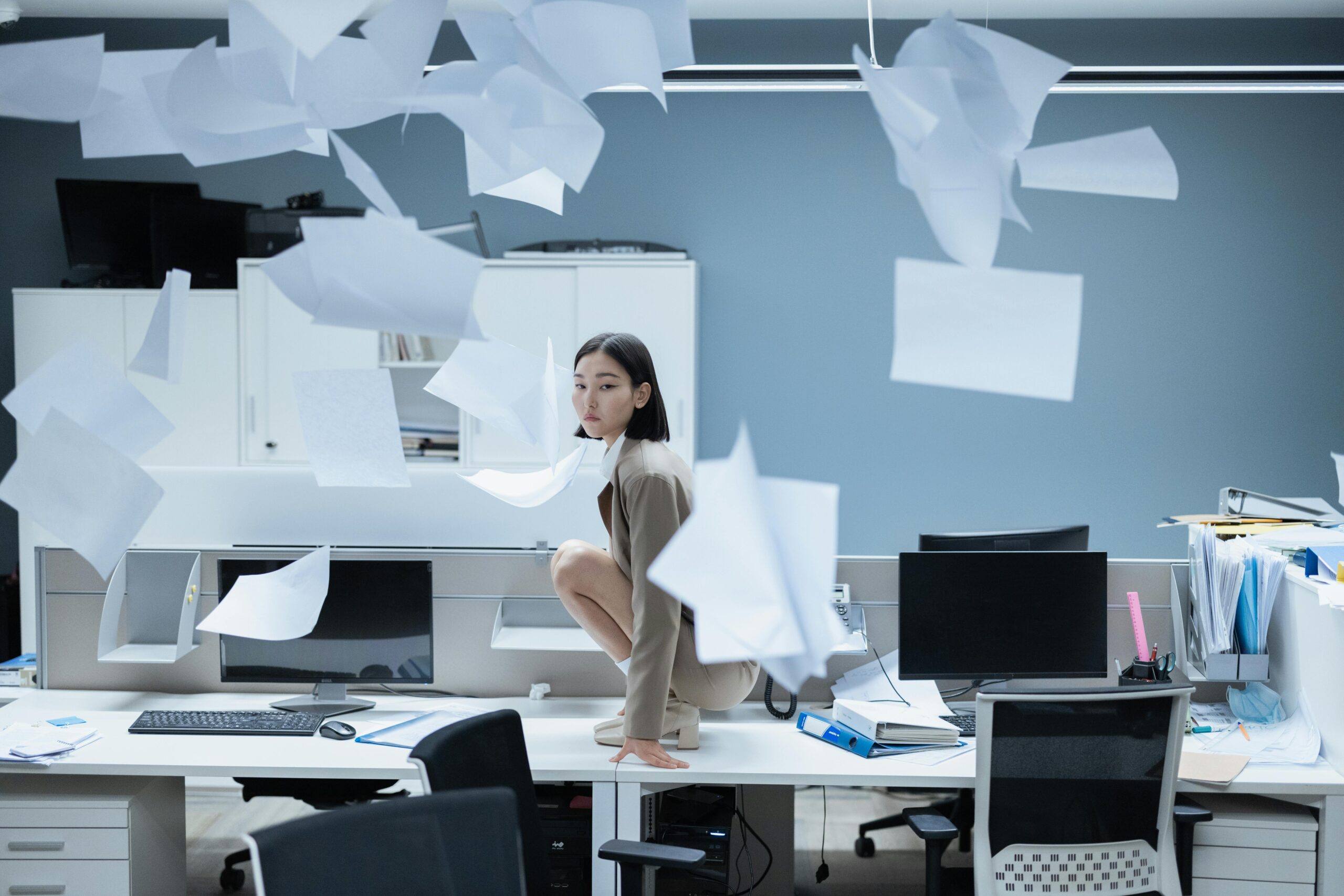 Woman Crouching on Desk Among Flying Papers in Office