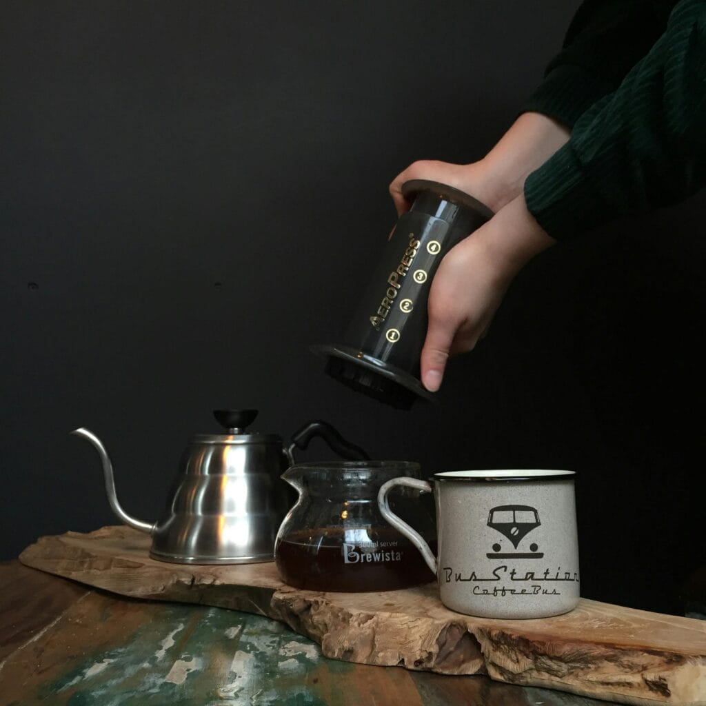 Hands brewing coffee with Aeropress beside kettle and mug on wooden table.