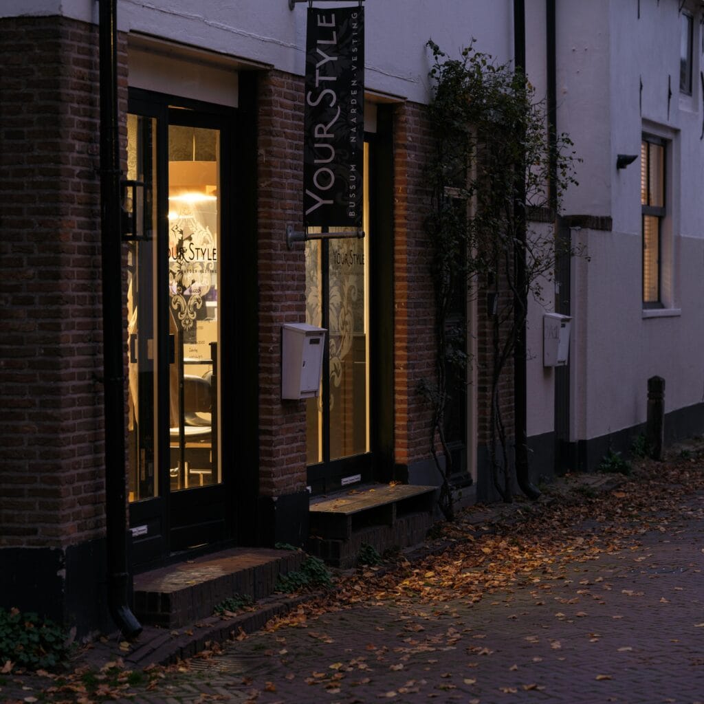 A warmly lit hair salon on a cobblestone street during dusk, autumn leaves scattered.